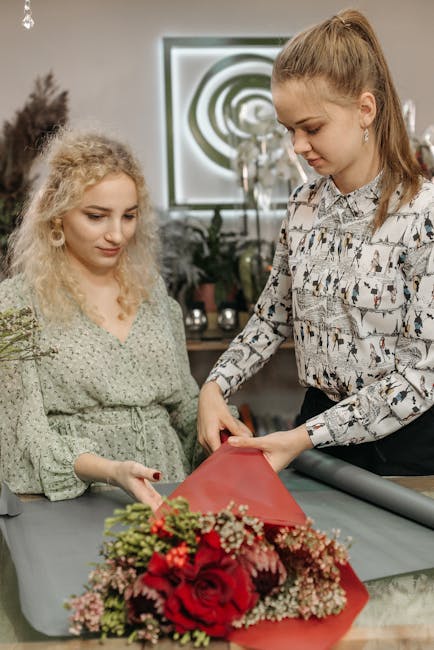 A floral arrangement featuring vibrant red roses, pink and white baby's breath, and greenery, being carefully wrapped in red tissue paper by a florist with light brown hair styled in a high ponytail, 