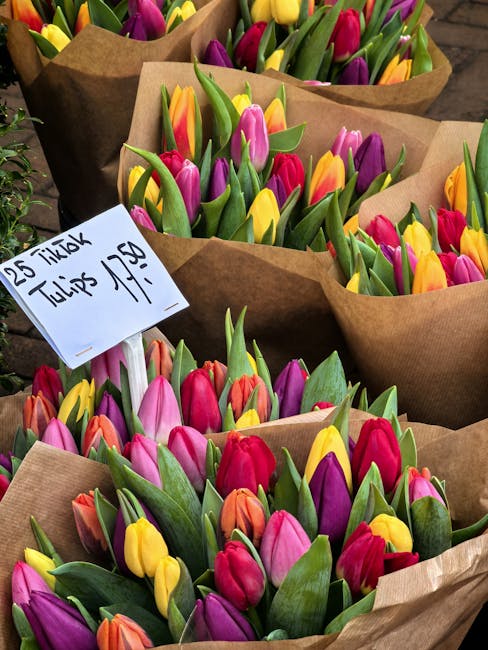 Multiple bouquets of fresh tulips arranged in brown kraft paper wrapping, showcasing vibrant colors including pink, purple, yellow, orange, and red. The tulips have green stems and leaves, appearing h