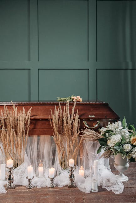 A floral arrangement featuring white roses, lilies, and greenery placed in a white vase situated on a wooden surface, partially obscured by tall, beige pampas grass stalks. The flowers are fresh, with
